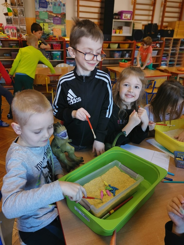 Dzieci odkrywają odszukują figurek dinozaurów w pojemniku z kaszą.