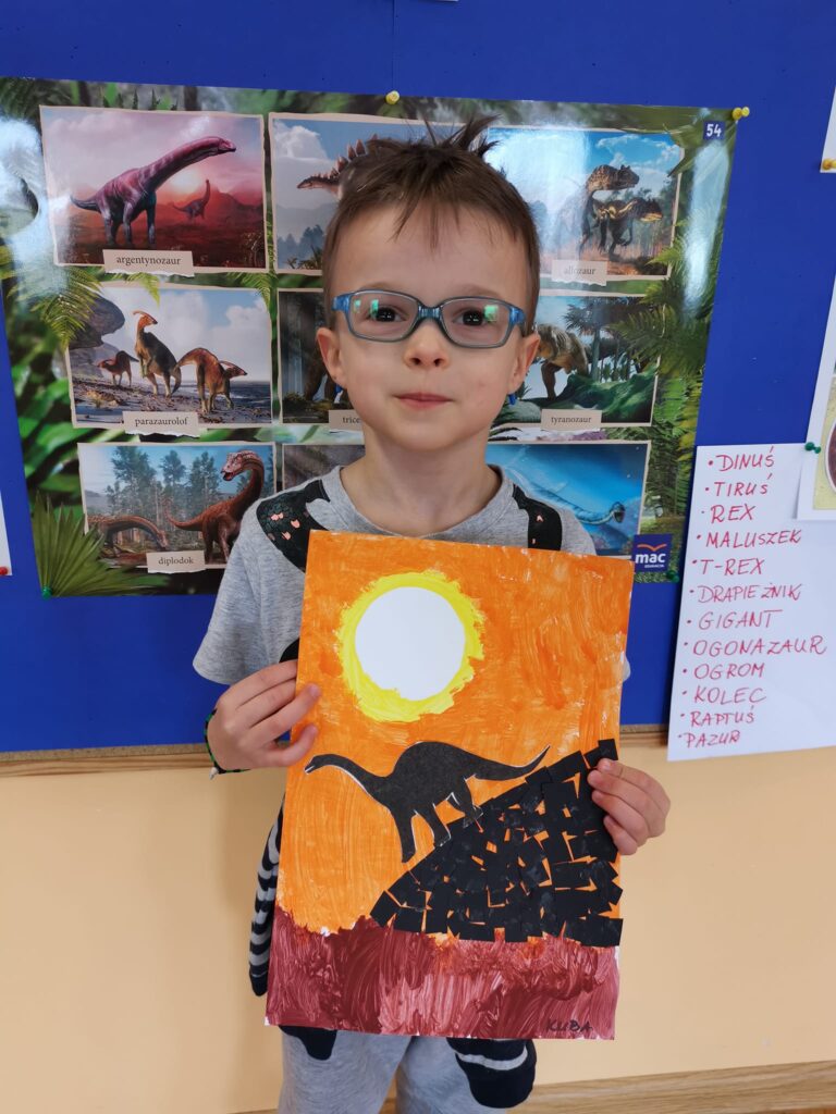 Chłopiec prezentuje swoją pracę plastyczną, dinozaur schodzący z góry przy zachodzie słońca.